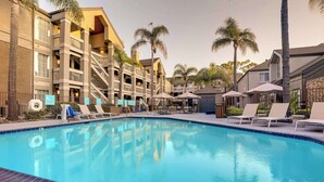 A swimming pool with lounge chairs and umbrellas, surrounded by multi-story buildings with balconies and palm trees.