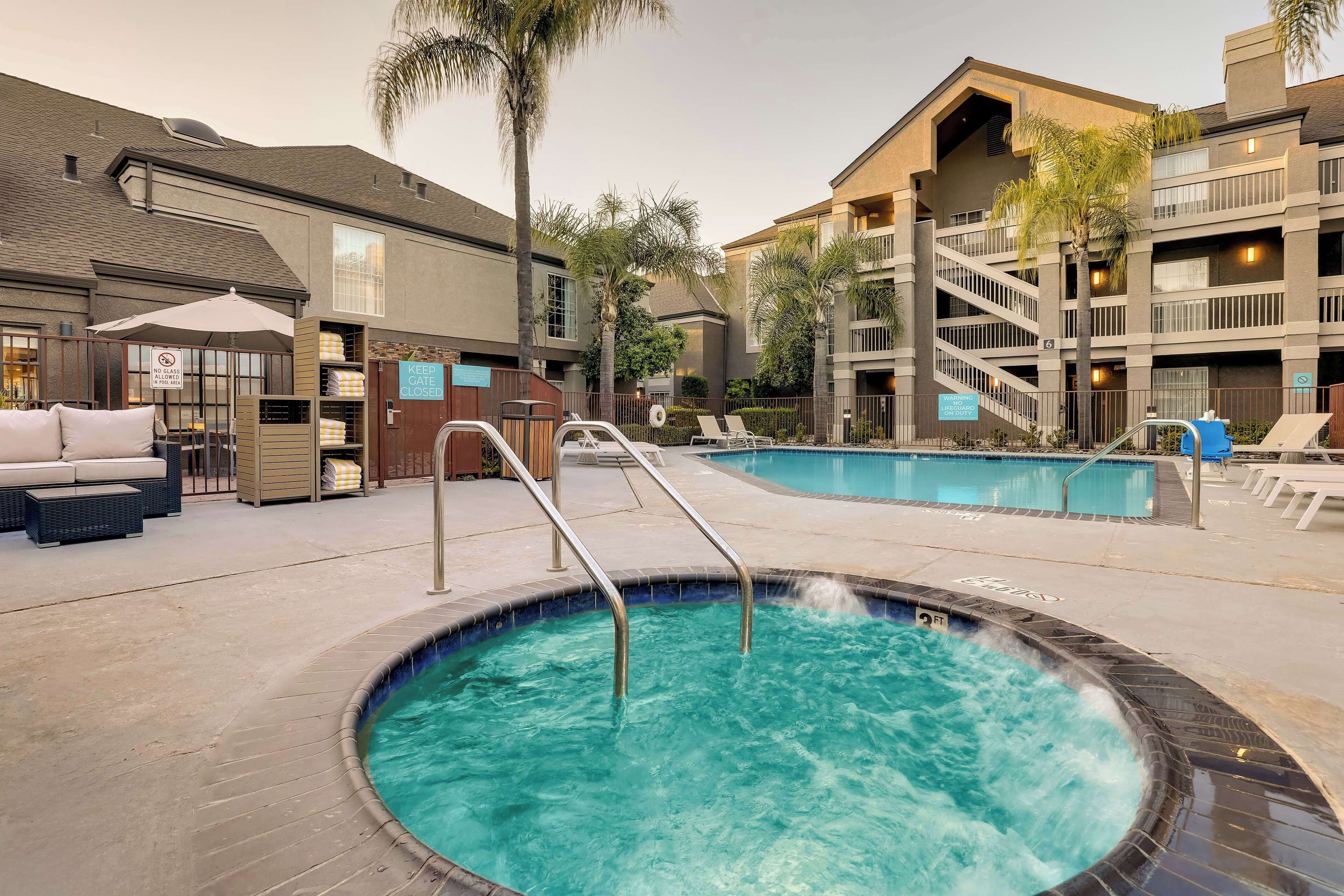 A hotel pool area with a hot tub, lounge chairs, and a staircase leading to an upper level.