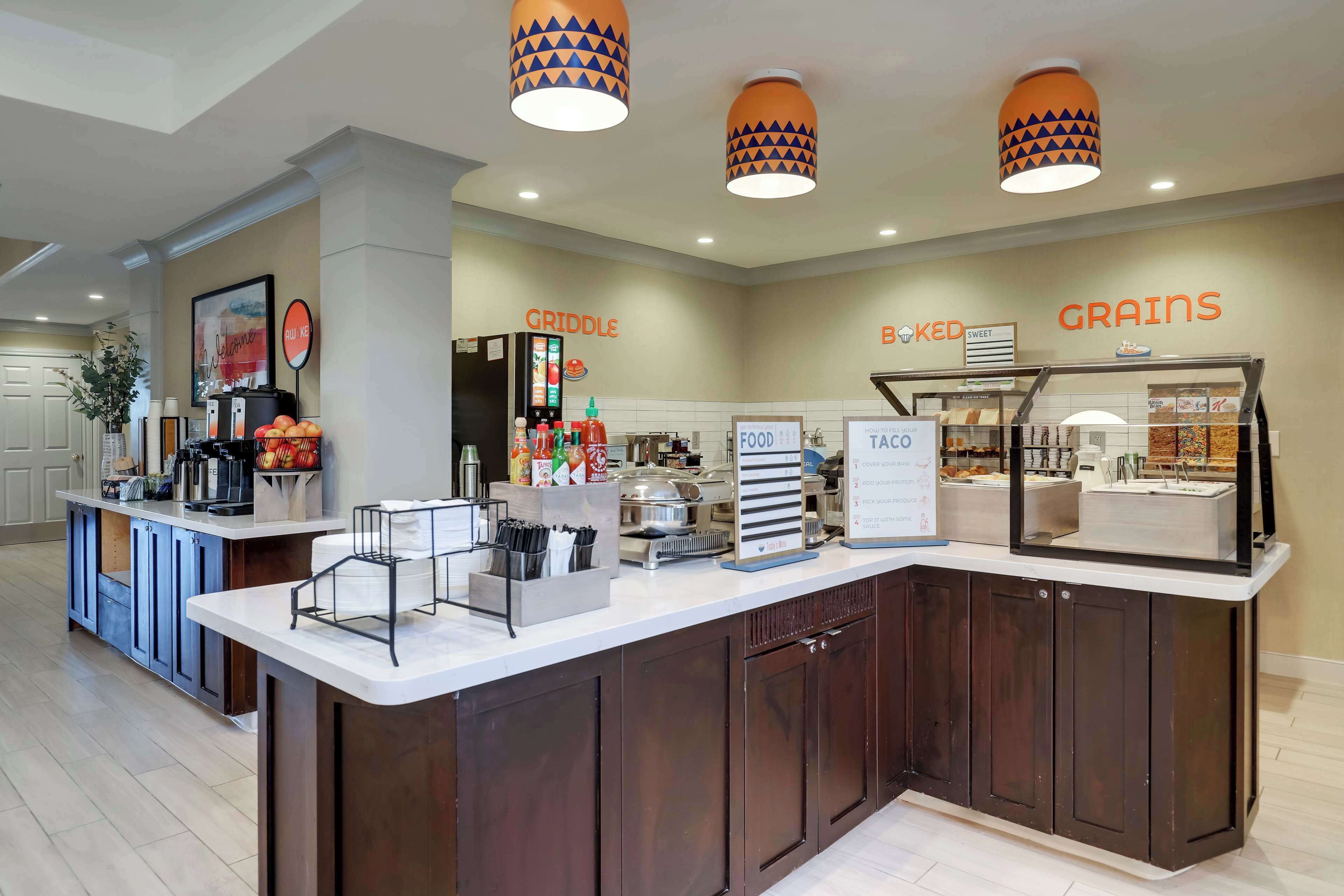 Zona de servicio de alimentos en un hotel, con un mostrador de autoservicio, una estación de condimentos y letreros que indican 'Griddle' y 'Baked Sweet Grains'.