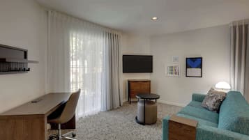 A modern hotel room with a wooden desk, a teal sofa, a TV mounted on the wall, and a small round table.