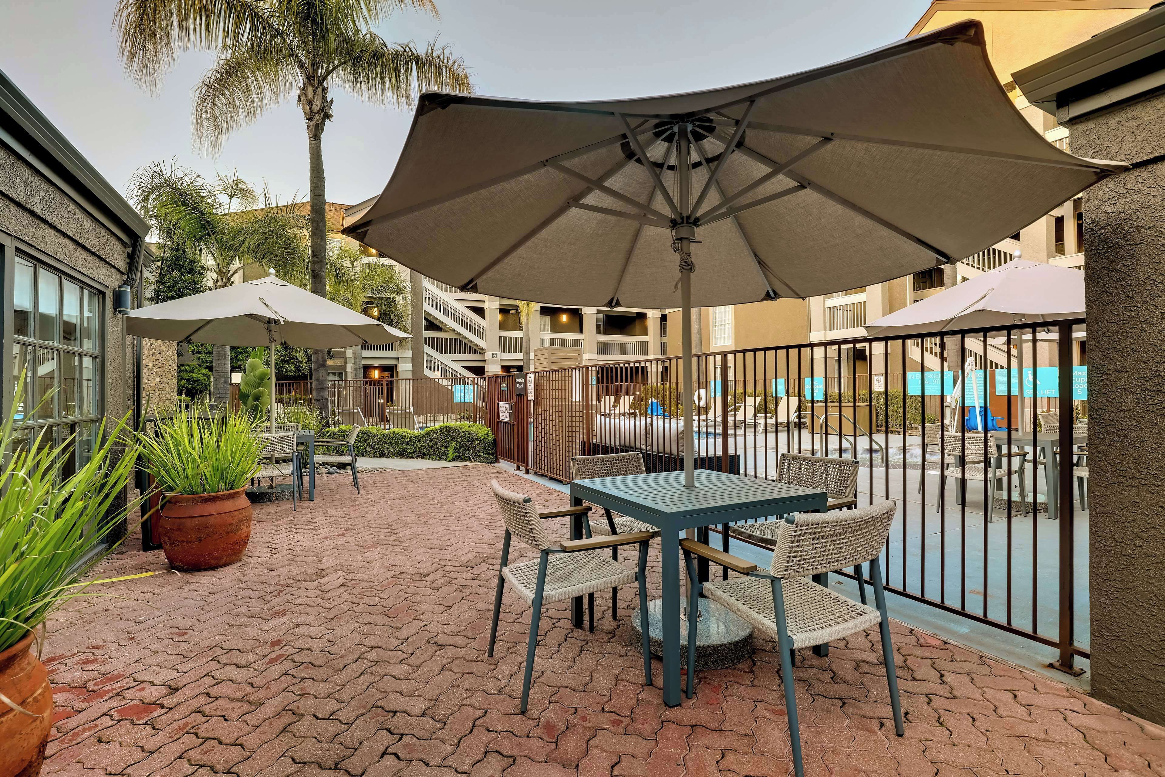A patio area with a table and chairs under a large umbrella, surrounded by potted plants and a brick floor.