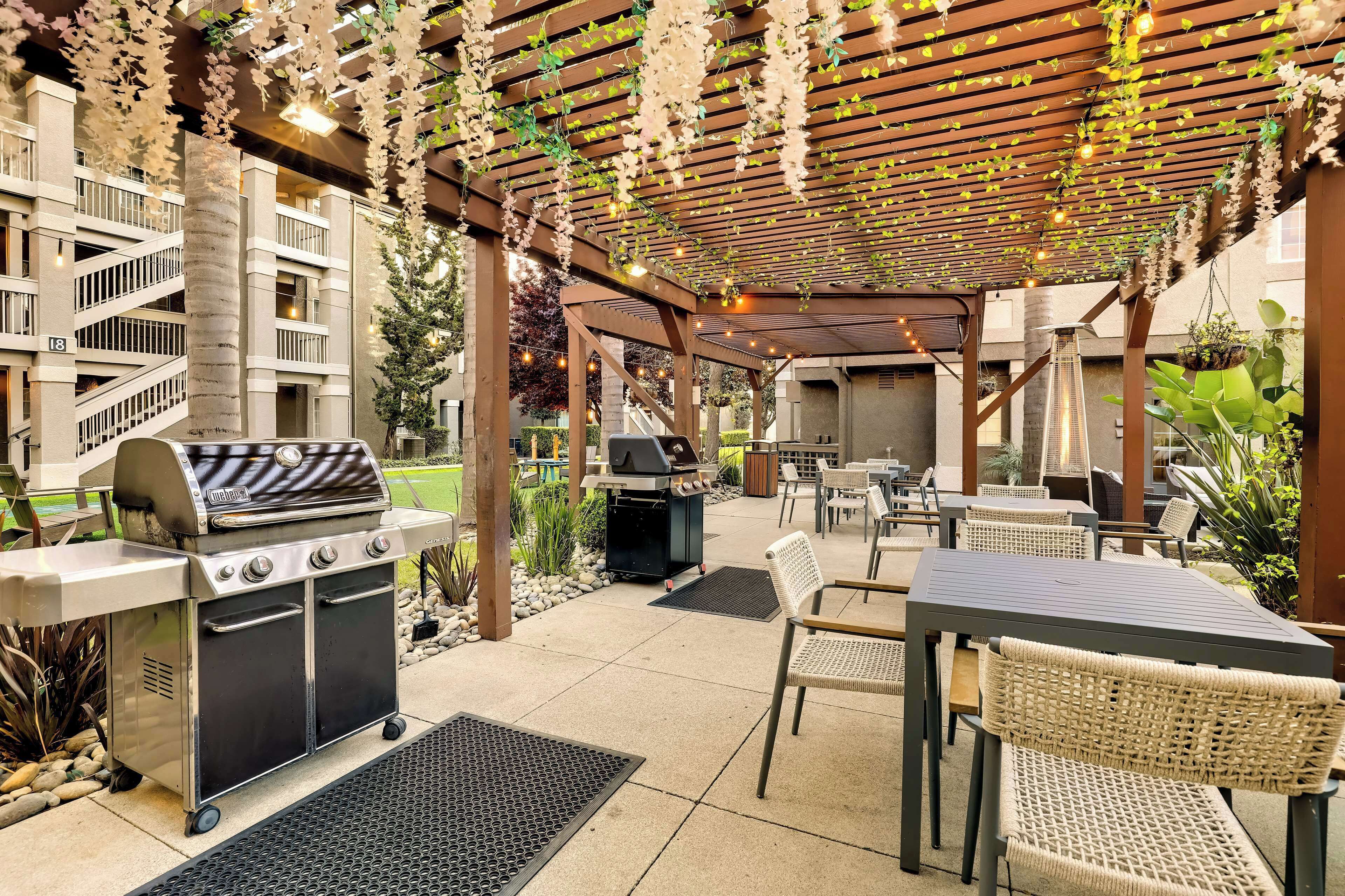 Zona de pati exterior amb una barbacoa, seients i una pèrgola amb plantes penjants.