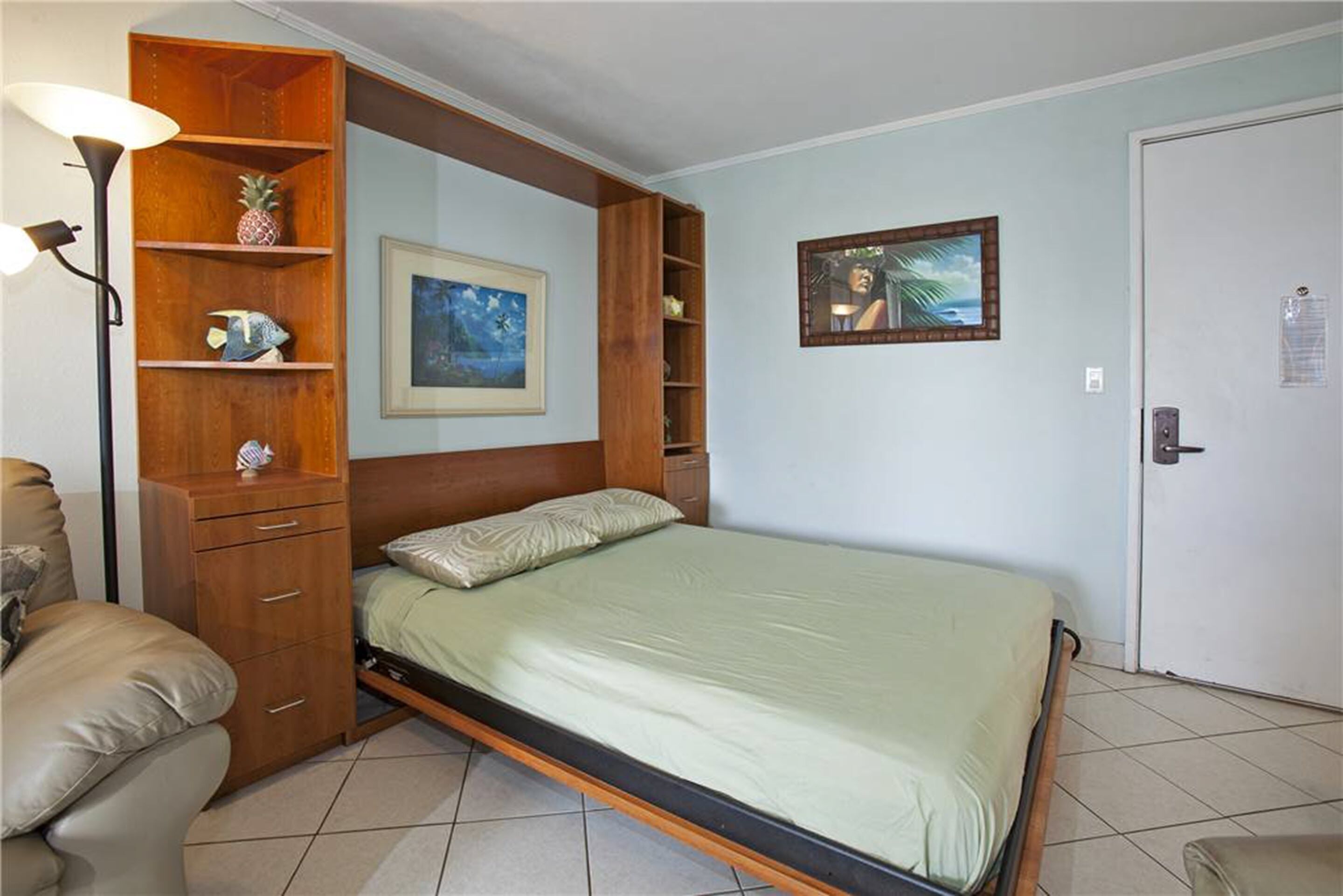 A single-bed room with a wooden headboard, a bedside lamp, a small shelf with a pineapple, and a framed picture on the wall.