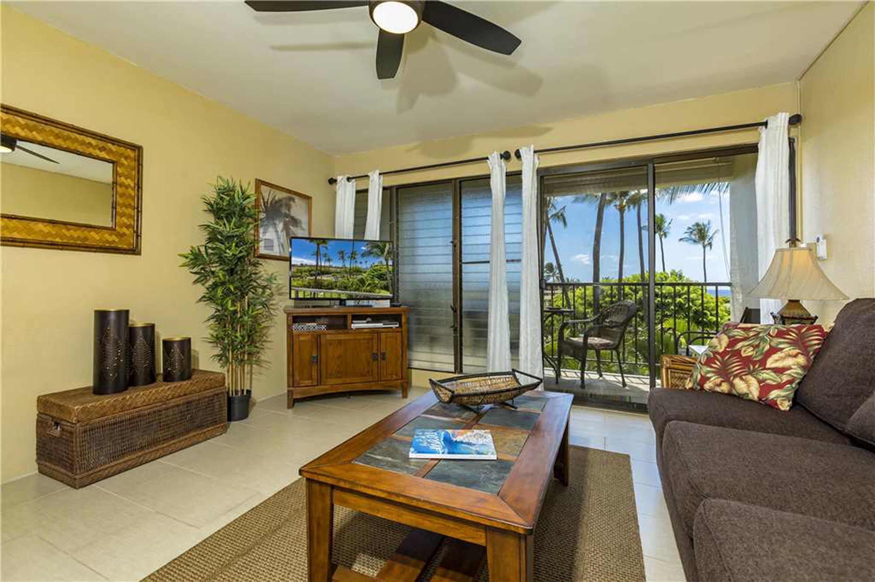 Un salón con sofá, mesa de centro y televisor sobre un mueble. Tiene un balcón con vistas a palmeras.