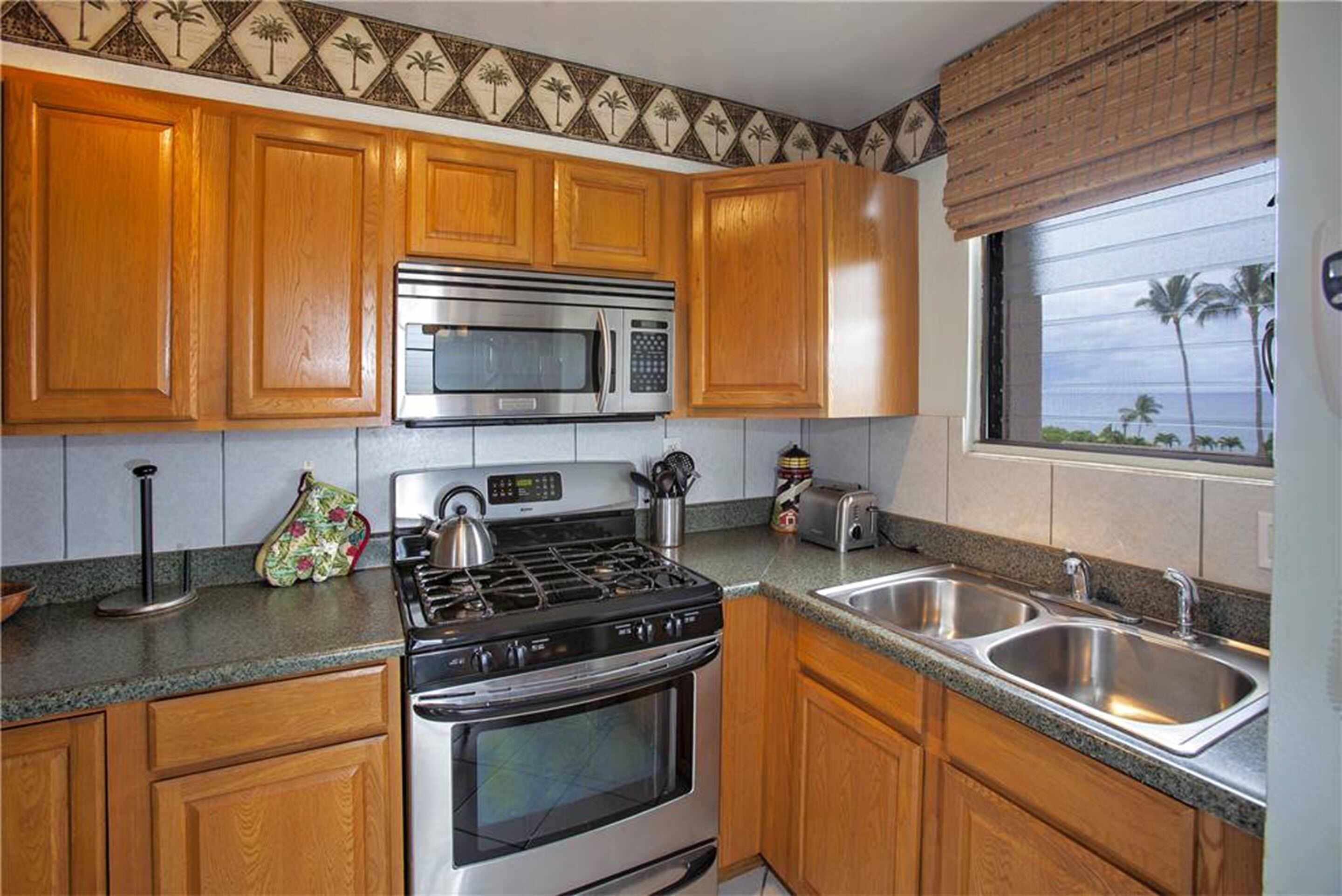 A kitchen with wooden cabinets, a stainless steel microwave, a gas stove, a toaster, and a window with a view of palm trees.