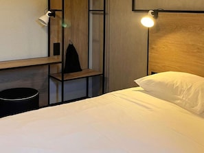 Ein Hotelzimmer mit Bett, Nachttisch, Lampe und einem Regal mit einer schwarzen Tasche.