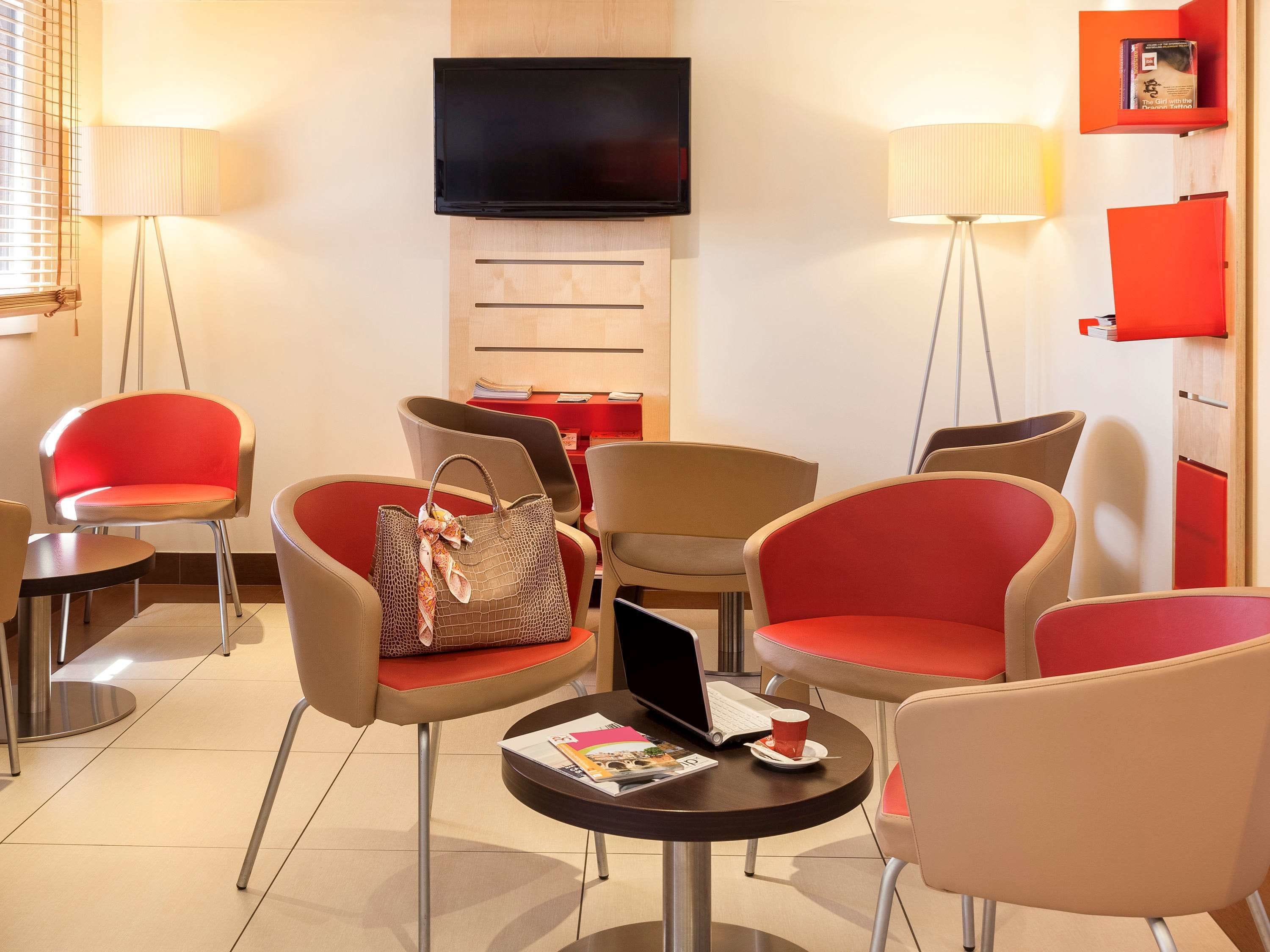 A modern hotel lobby with red and beige chairs, a flat-screen TV, and a red bookshelf.