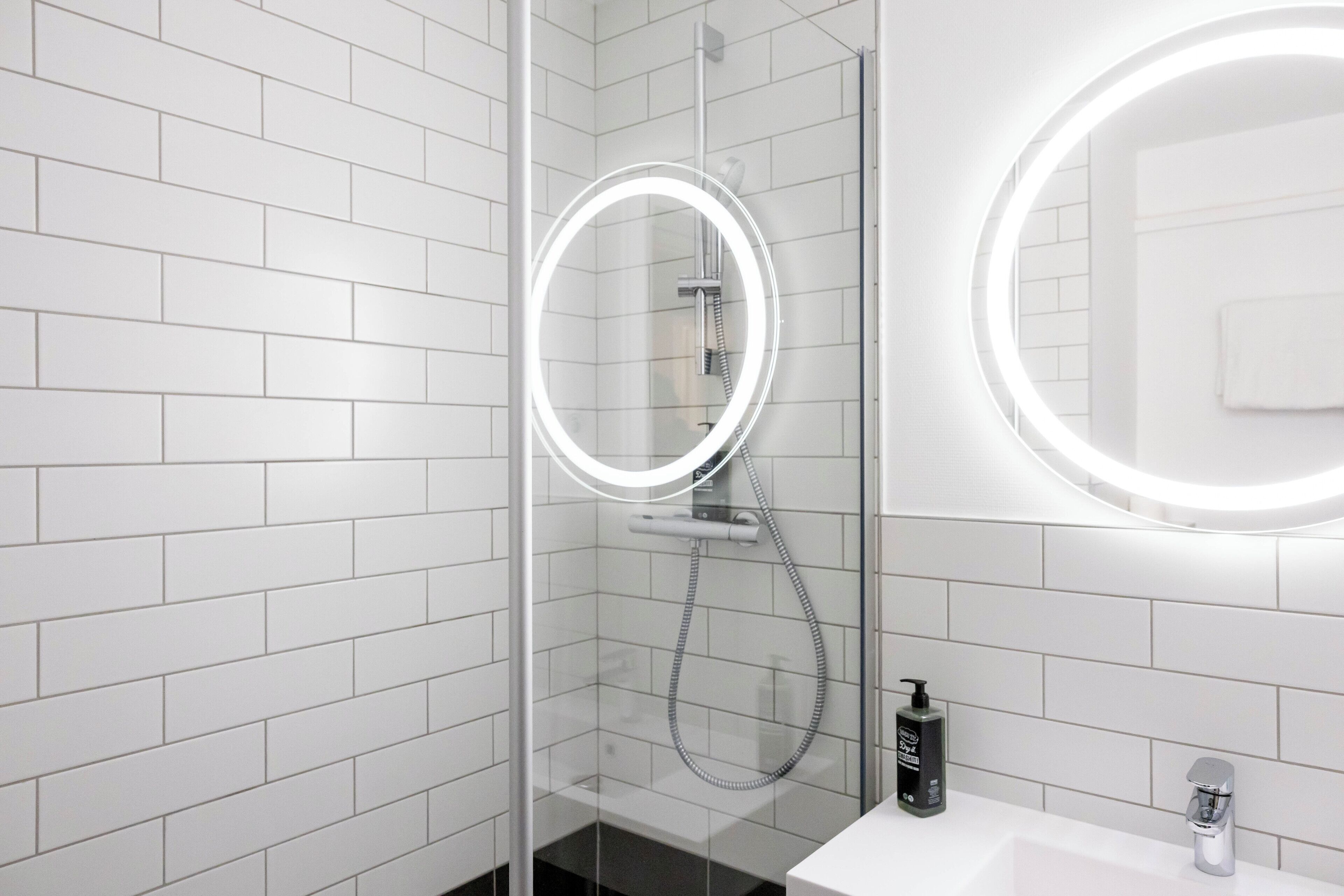 A modern bathroom with white subway tiles, a glass-enclosed shower with a handheld showerhead, and a round mirror with a lighted frame.
