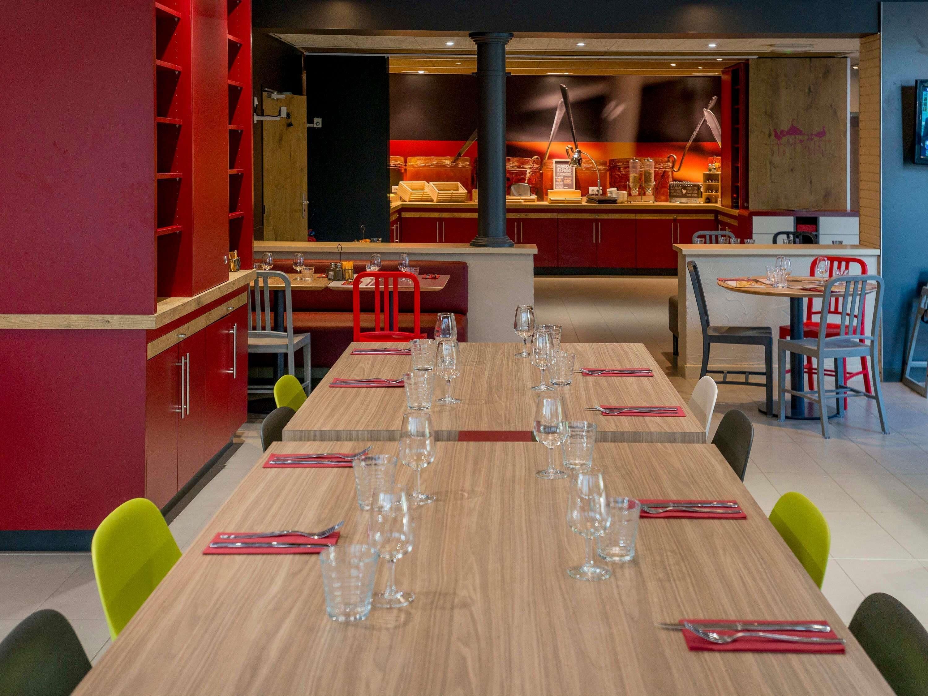 Een restaurant met een lange houten tafel gedekt voor een maaltijd, rode stoelen en een toonbank met een presentatie van etenswaren.