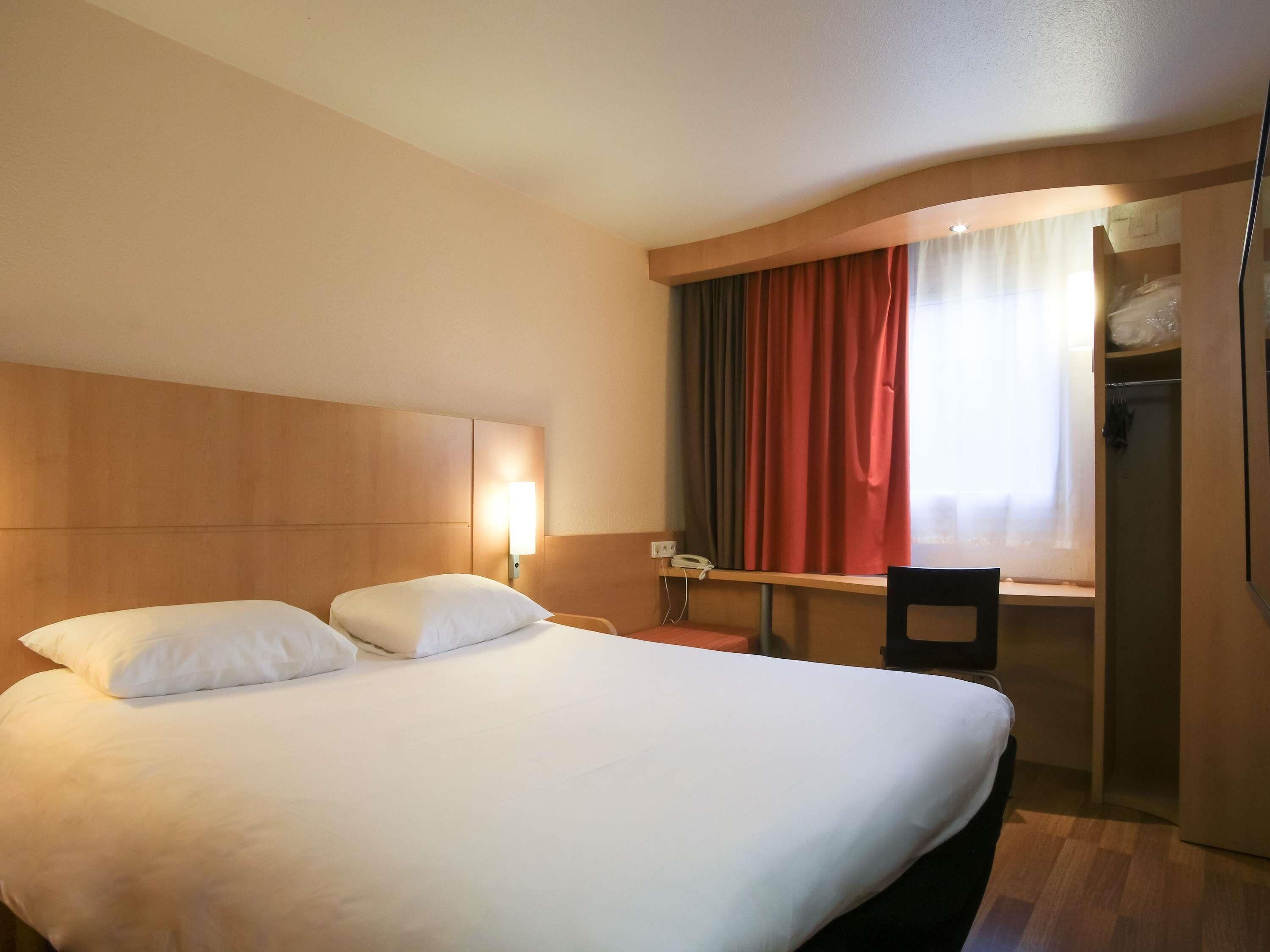 Een hotelkamer met een groot bed, een bureau en een stoel. Er is een raam met gordijnen, een telefoon op het bureau en een kledingkast.