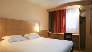 Een hotelkamer met een groot bed, een bureau en een stoel. Er is een raam met gordijnen, een telefoon op het bureau en een kledingkast.