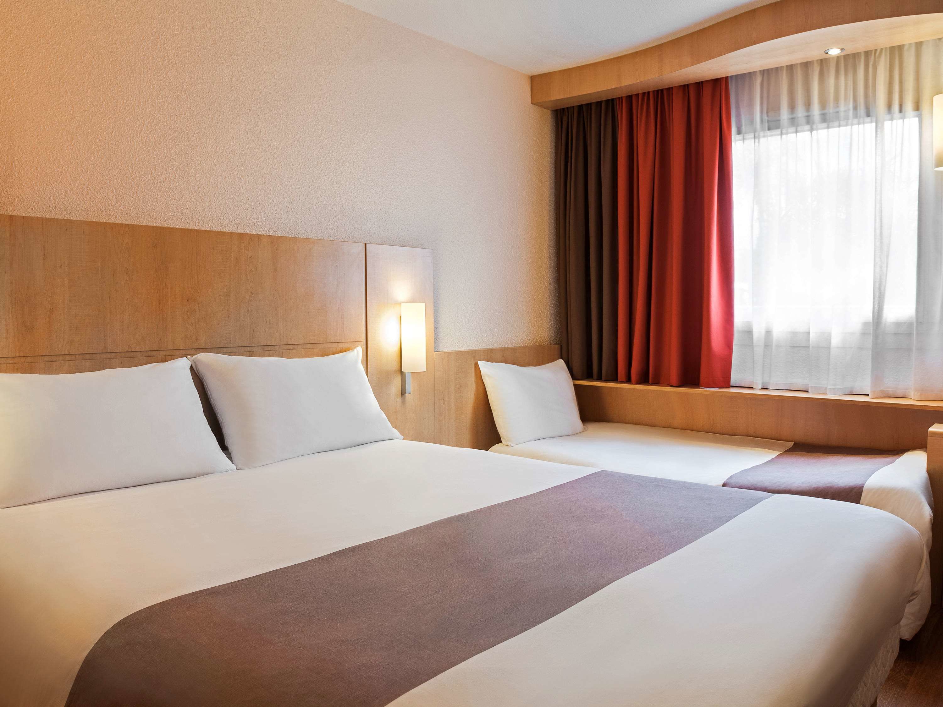 A hotel room with two beds, a wooden headboard, a window with red curtains, and a wall-mounted light.