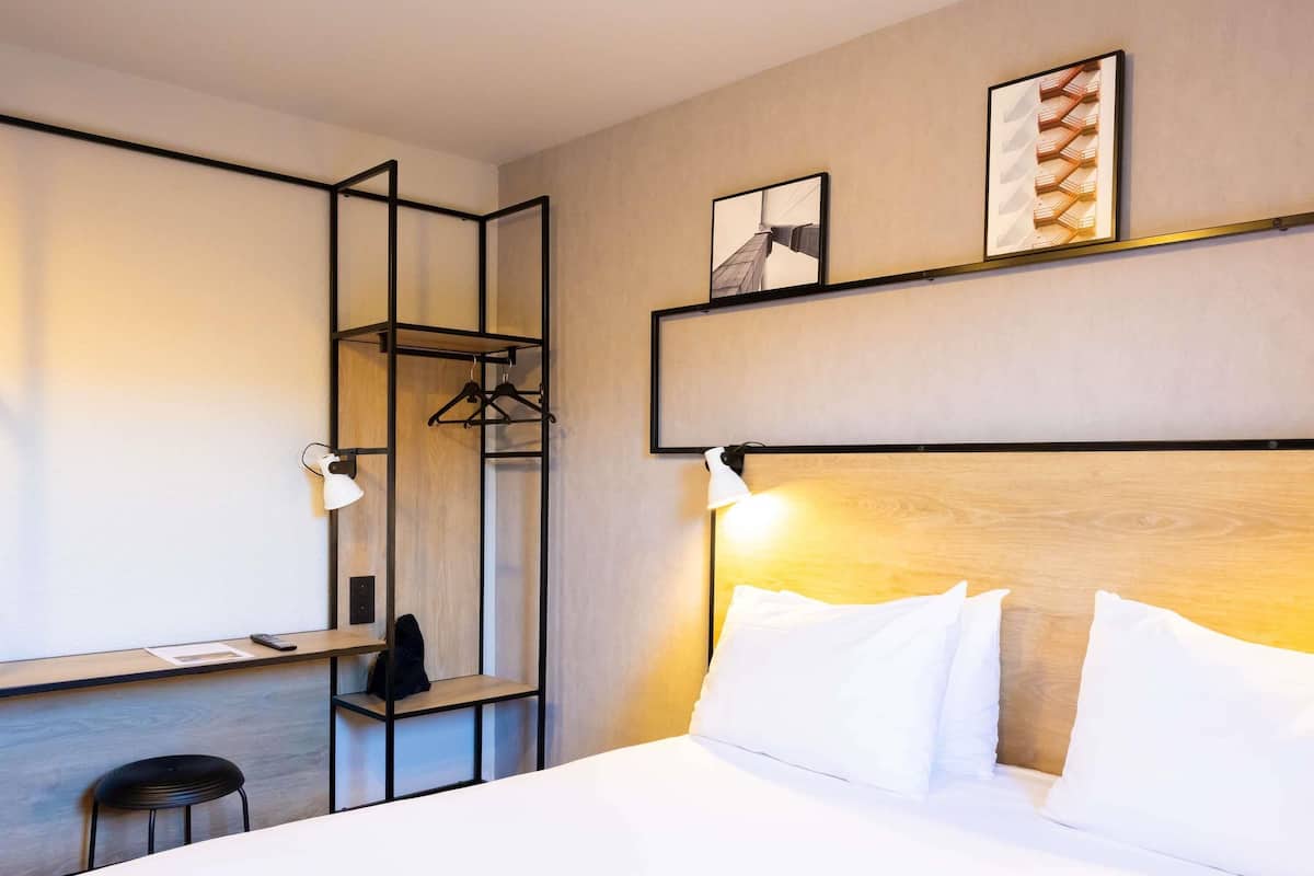 Habitación de hotel con cama, escritorio, silla, perchero y dos lámparas fijadas a la pared.