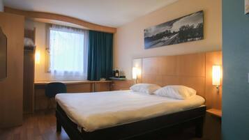 A hotel room with a bed, a desk, a chair, a window with curtains, a painting on the wall, and bedside lamps.