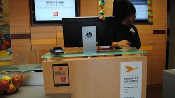 A hotel reception desk with a computer, a priority welcome sign, and a screen displaying 'BIENVENUE À L'HÔTEL IBIS TOULOUSE BLAGNAC AEROPORT'.