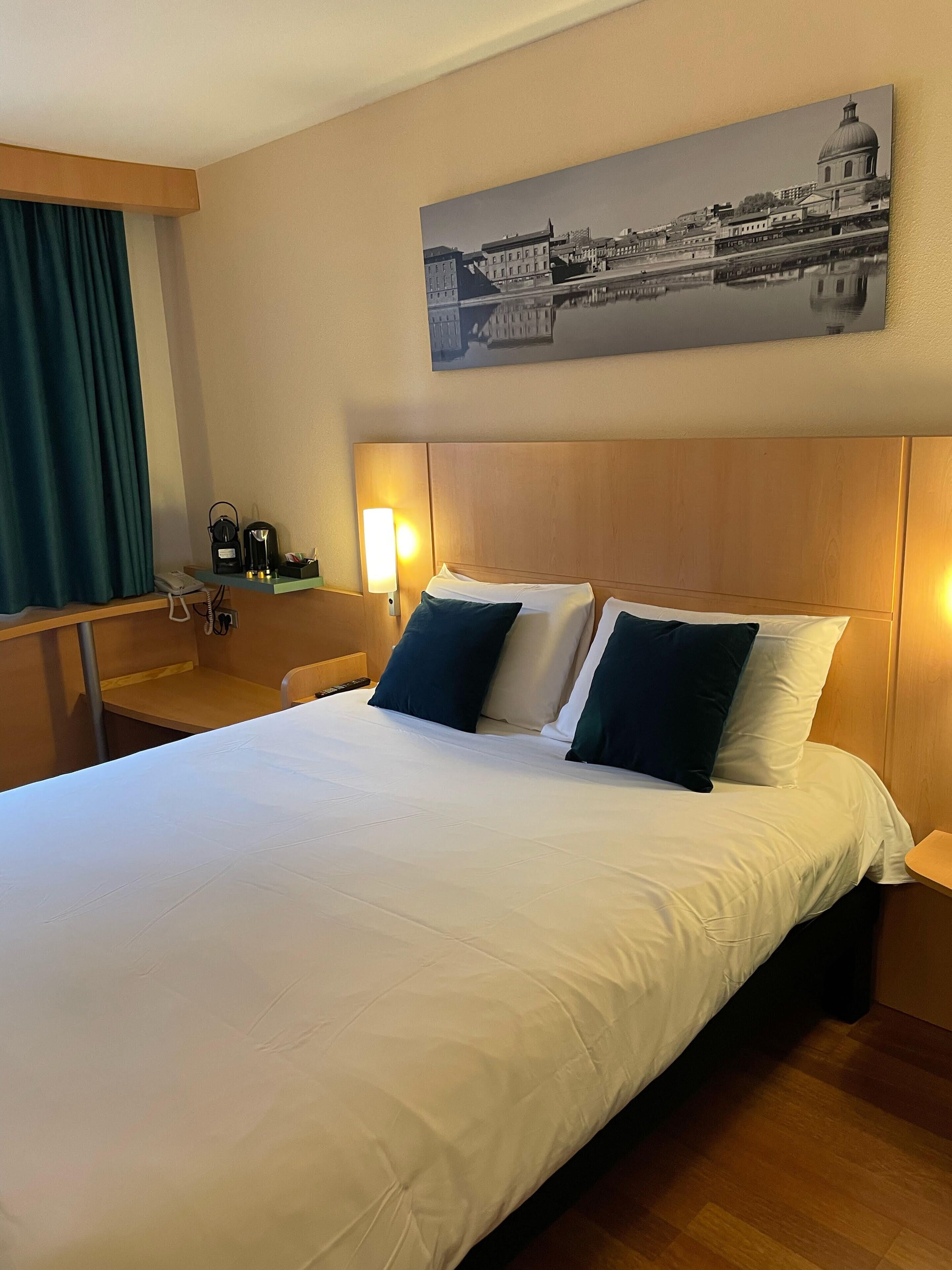 A hotel room with a large bed, a desk with a phone, a coffee maker, and a wall-mounted lamp.