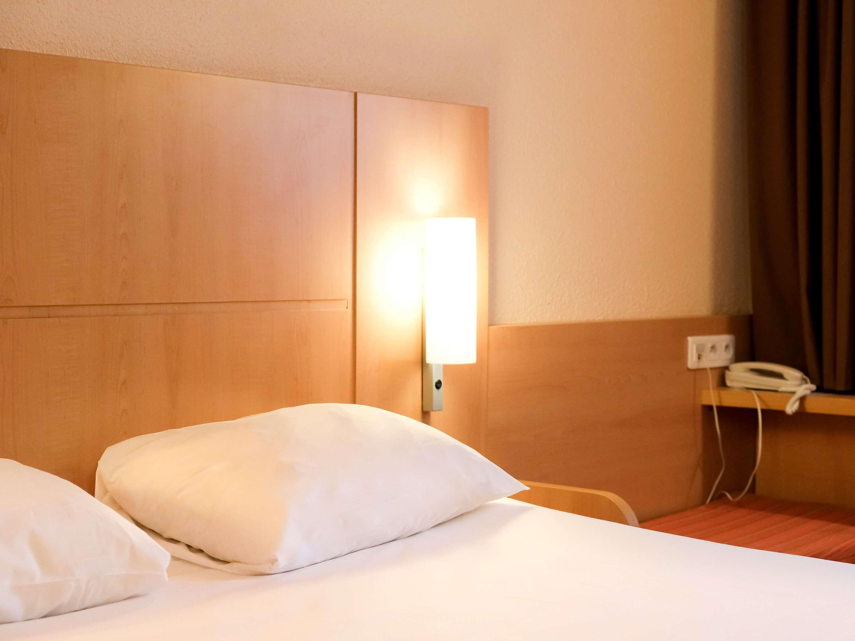 Quarto de hotel com cabeceira de madeira, luminária de mesa, telefone e cama com roupa de cama branca.
