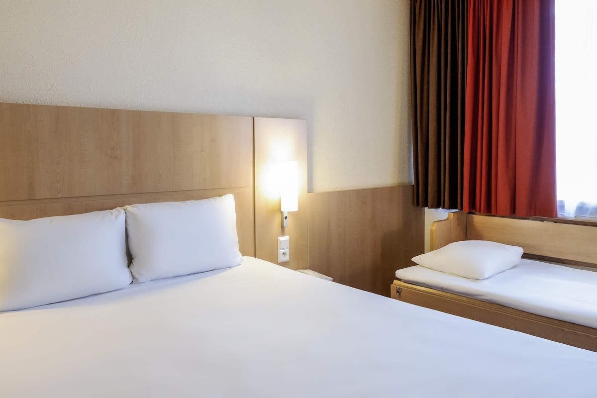 Una habitación de hotel con una cama grande, una cama más pequeña, una mesita de noche con lámpara y un ventanal con cortinas.