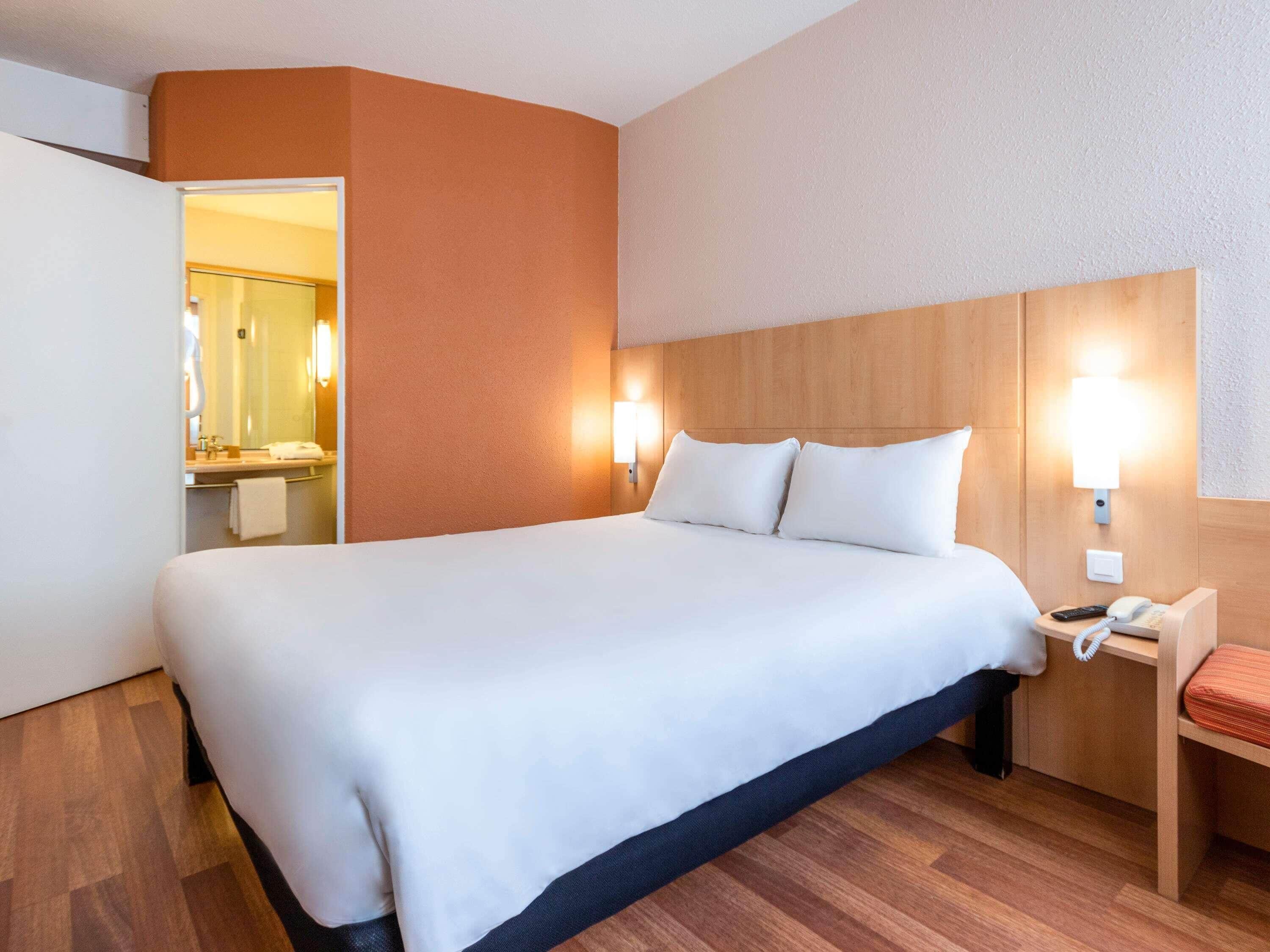 Hotelkamer met een groot bed, houten hoofdeinde, nachtlampjes, een nachtkastje met telefoon, en een badkamer zichtbaar door een open deur.