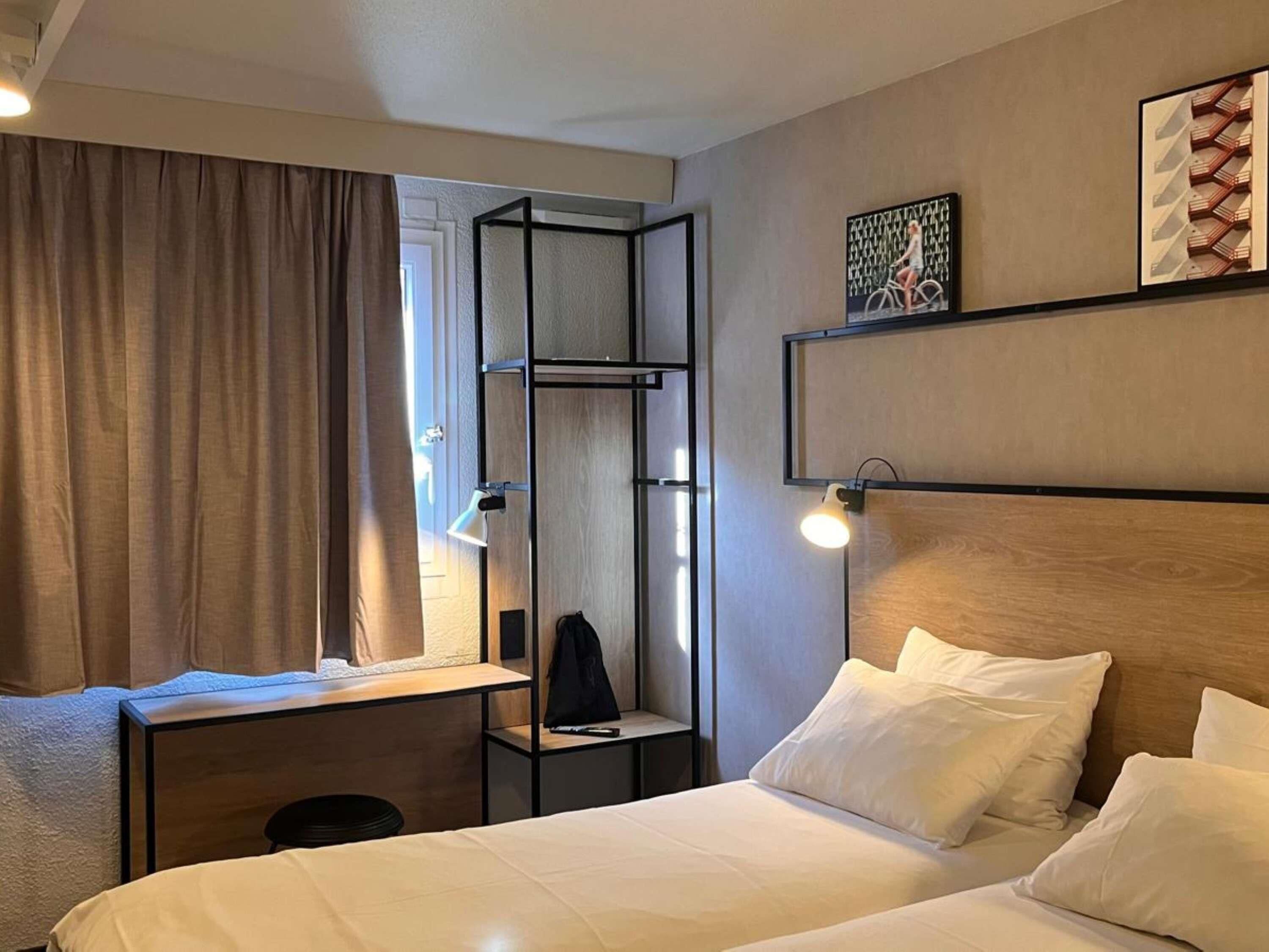 Une chambre d’hôtel comprenant un lit, un bureau avec une chaise, une armoire, une étagère avec des œuvres d’art et une fenêtre avec des rideaux.