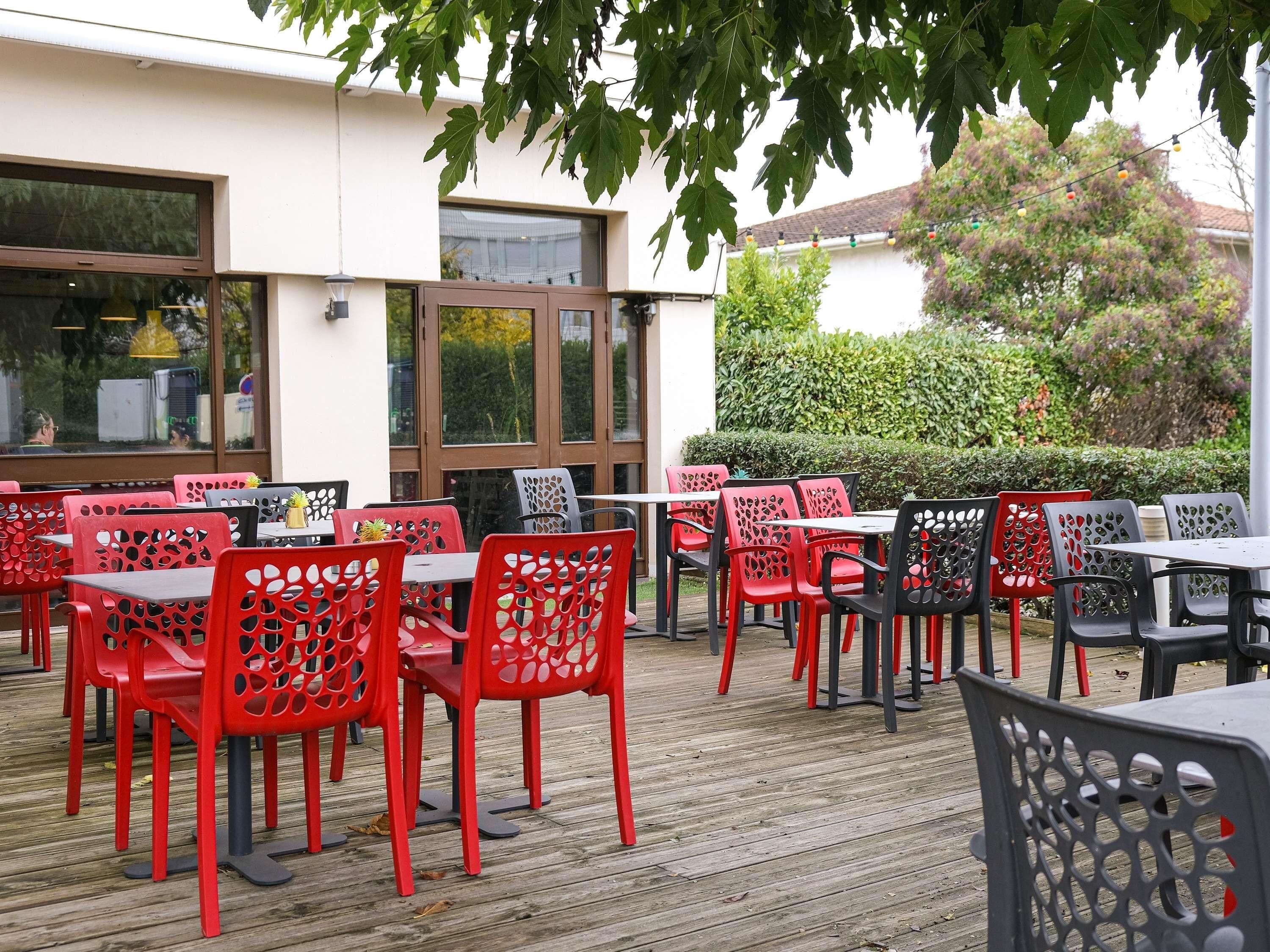 An outdoor seating area with red and black chairs and tables, large windows, and a view of a building and trees.