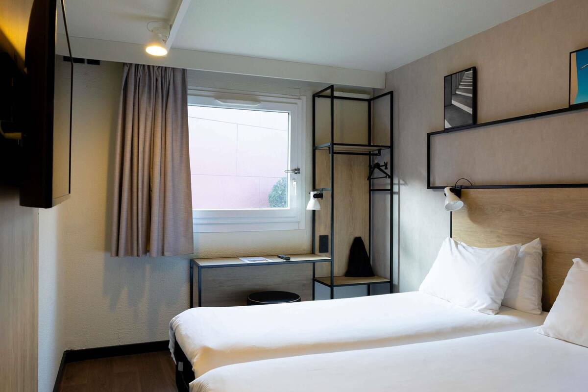 Habitación de hotel con dos camas, un escritorio, un televisor, una ventana con cortinas y una estantería con lámpara.
