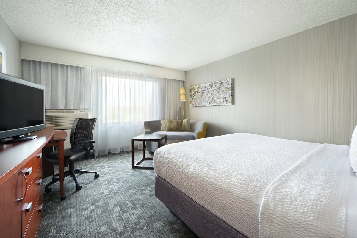 A hotel room with a bed, a desk with a television, a chair, a small table, and a window with curtains.