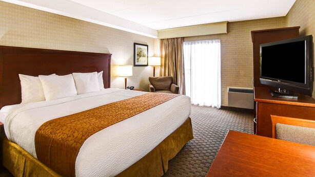 A hotel room with a large bed, a desk with a television, a chair, and a window with curtains. - Quality Inn (Kitchener)