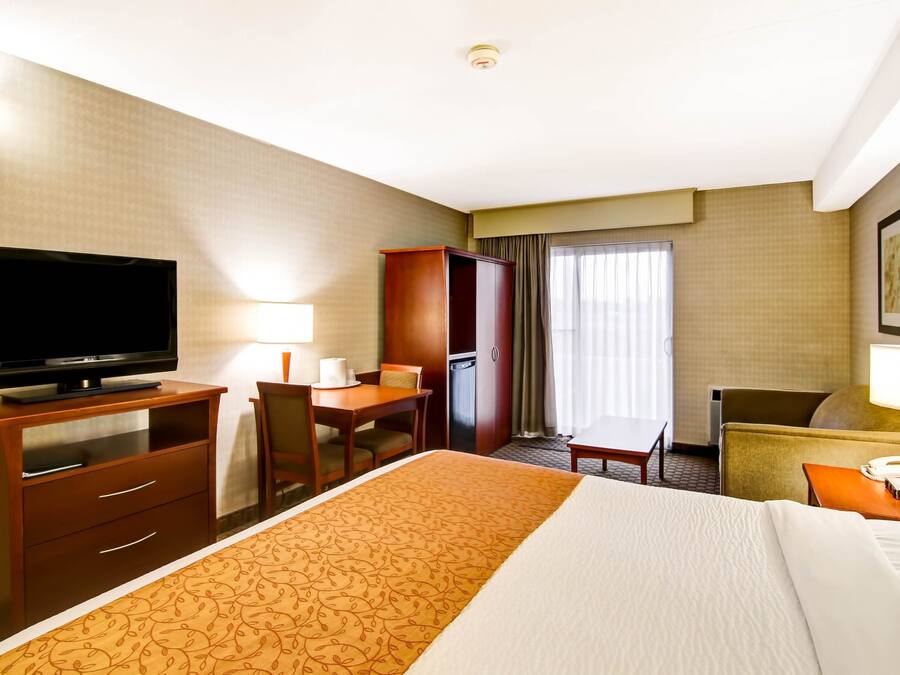 Quarto de hotel com cama, televisão, escrivaninha, cadeira, guarda-roupa e janela com cortinas.