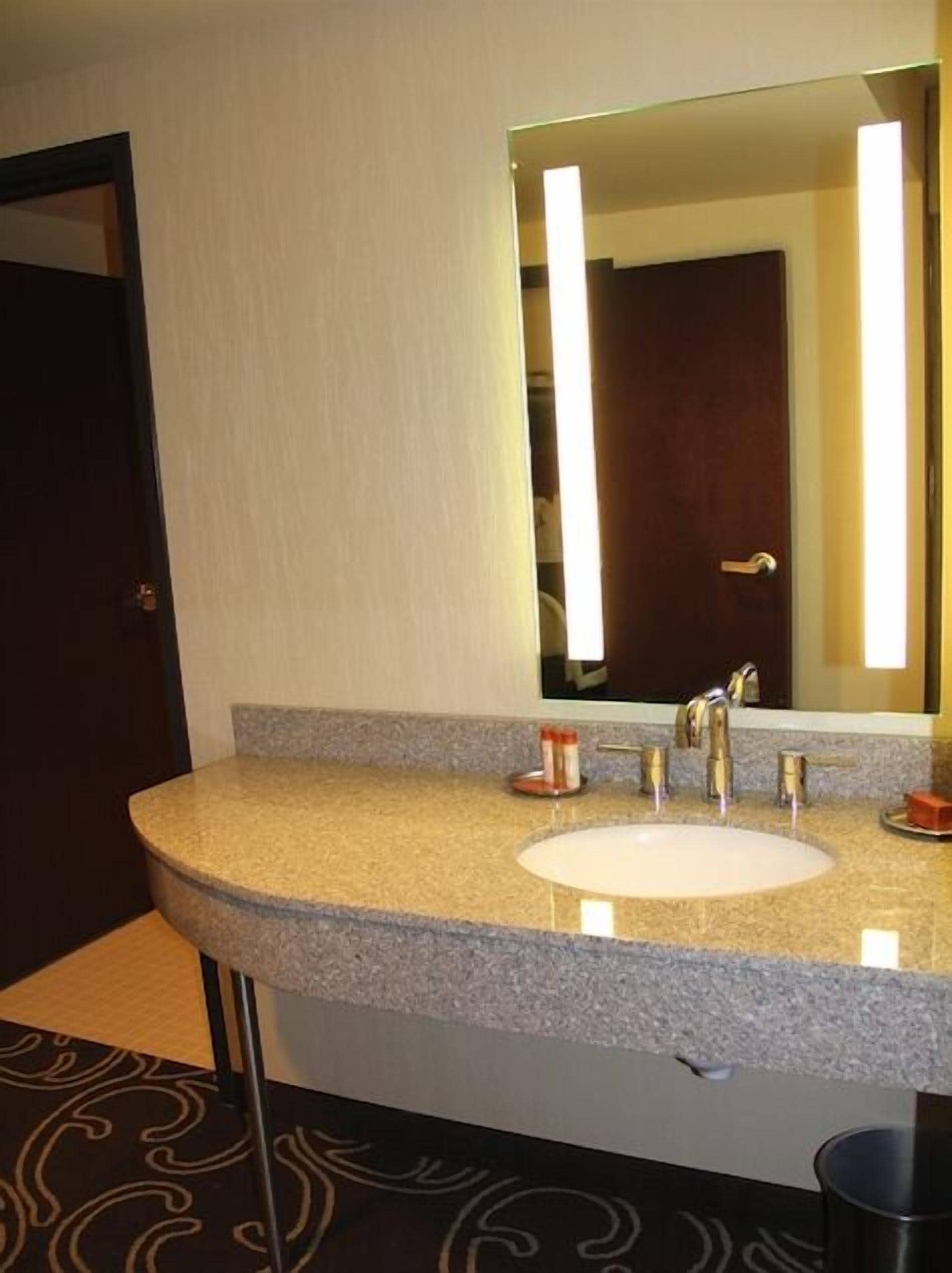 A hotel bathroom with a granite countertop sink, a large mirror, and a dark door.