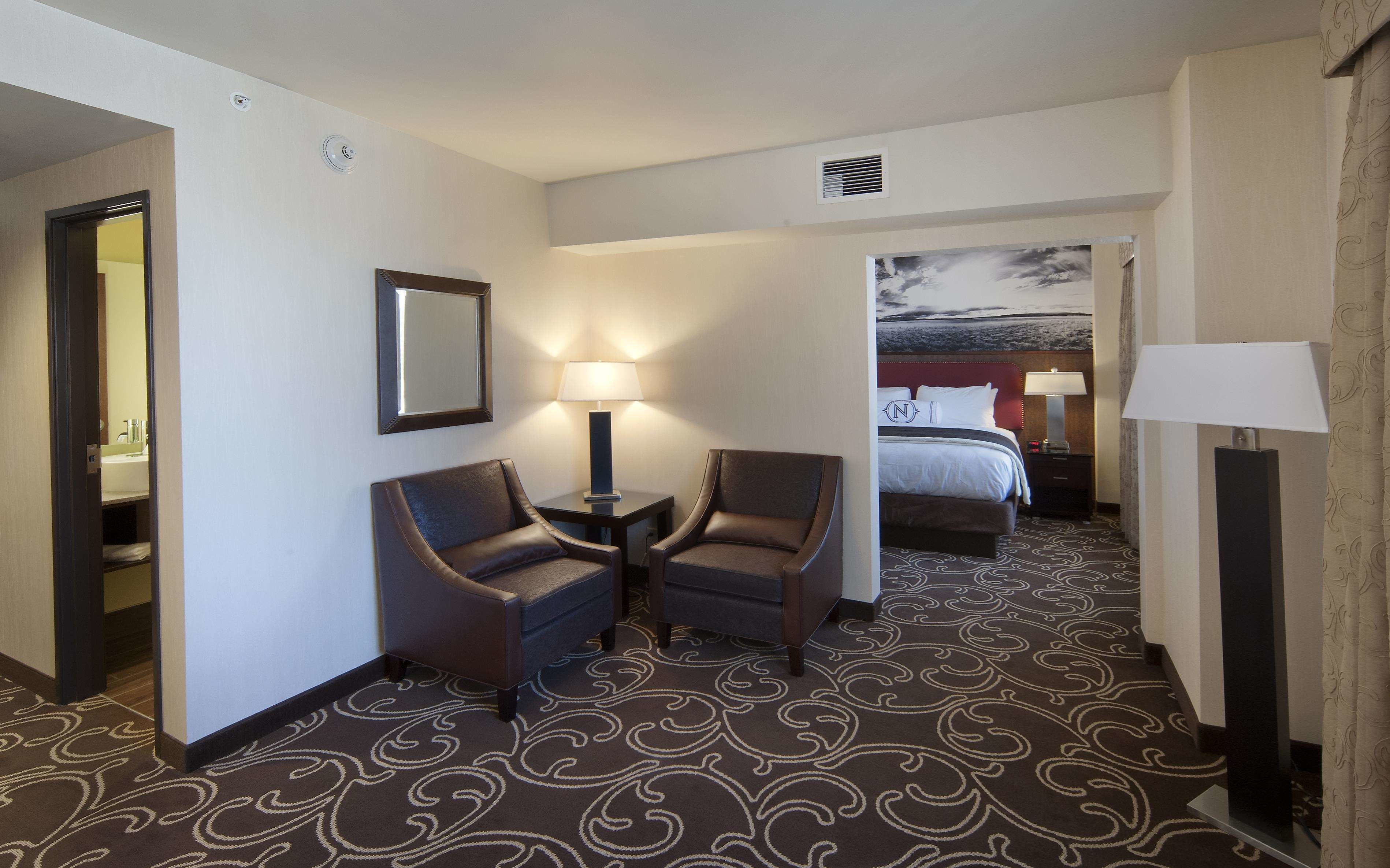 A hotel room with a bed, two armchairs, a nightstand, a lamp, and a patterned carpet.