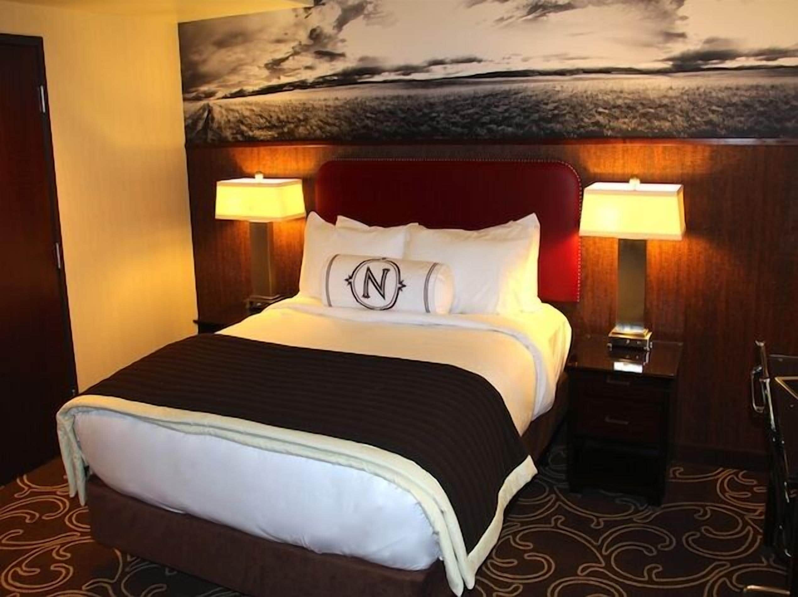 Ein Hotelzimmer mit einem großen Bett, zwei Nachttischlampen, einem Kopfteil mit dem 'N'-Logo und einem Wandgemälde.