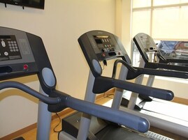 Three stationary bikes in a gym with control panels, a window, and a television on the wall.