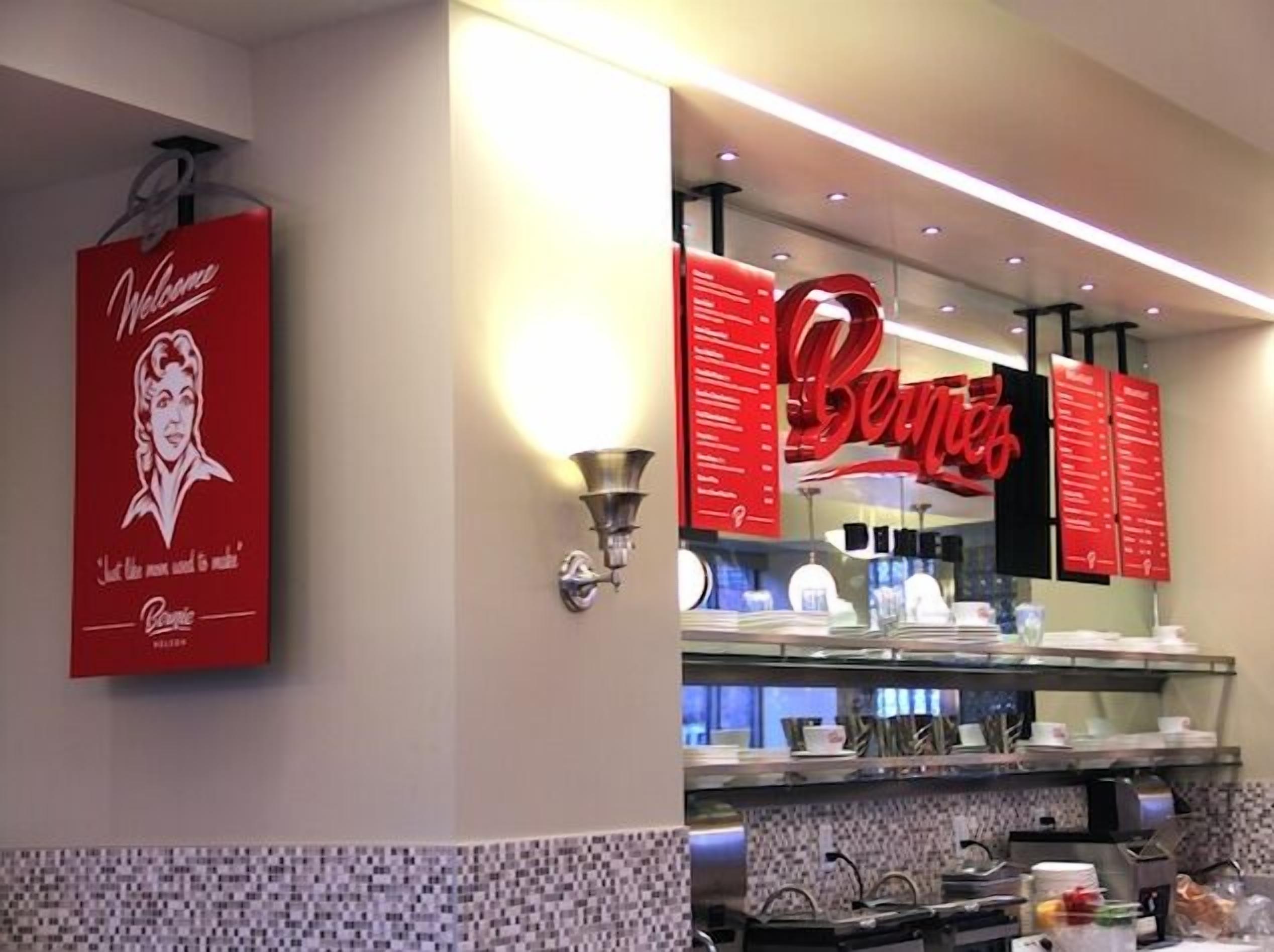 Interior de restaurante com um letreiro vermelho indicando "Welcome" e "Coca-Cola" na parede, um revestimento de azulejos e um balcão com diversos pratos e utensílios.