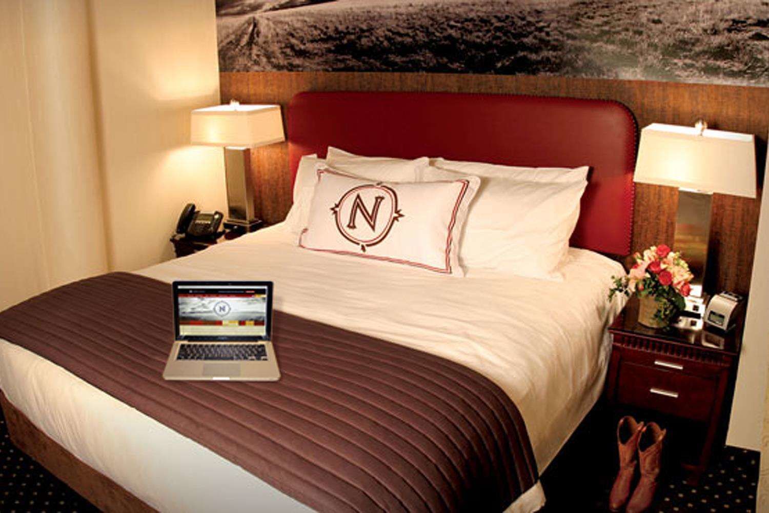 A hotel room with a bed, a laptop on the bed, a nightstand with a lamp, a phone, and a small table with a vase of flowers.