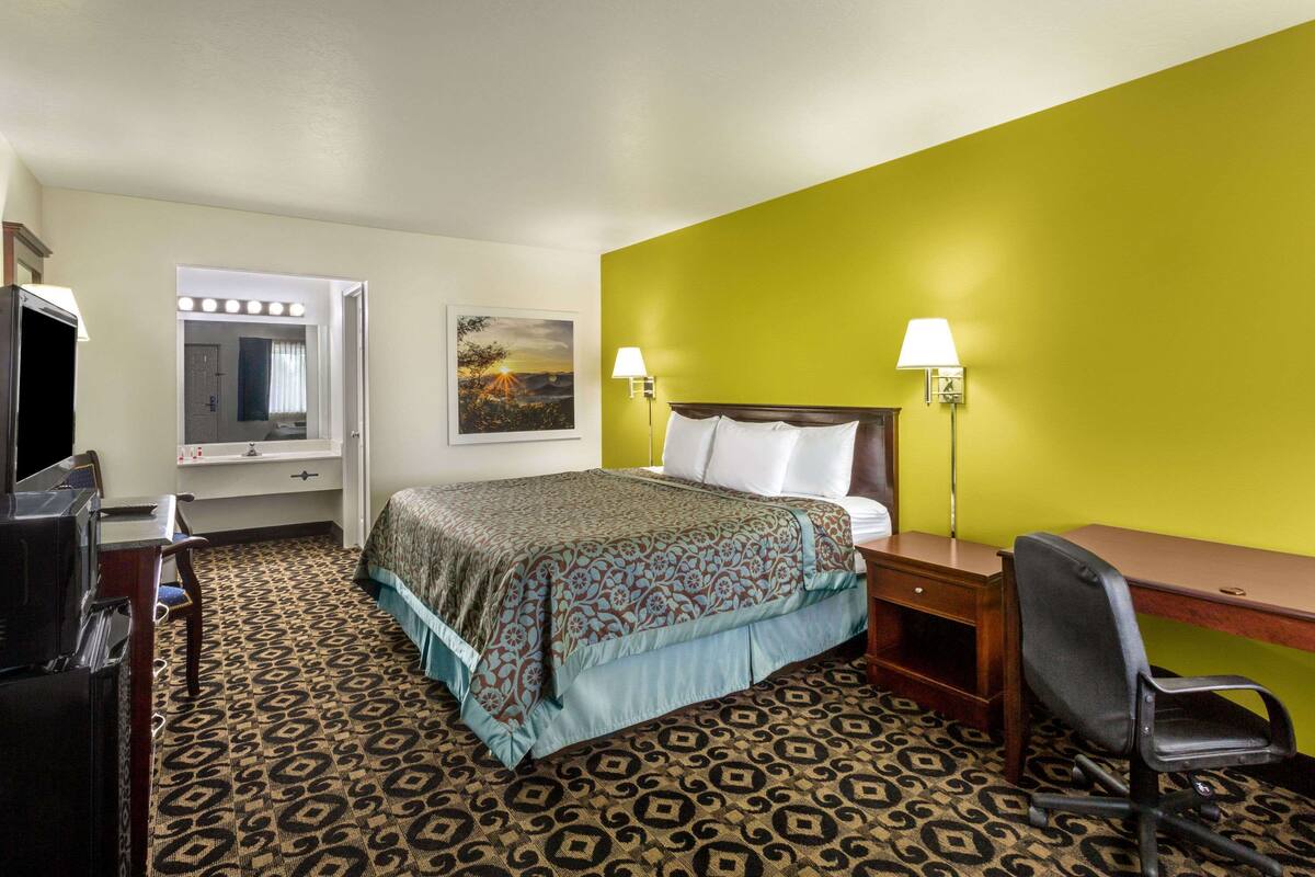 A hotel room with a bed, desk, chair, and a mirror with lights.