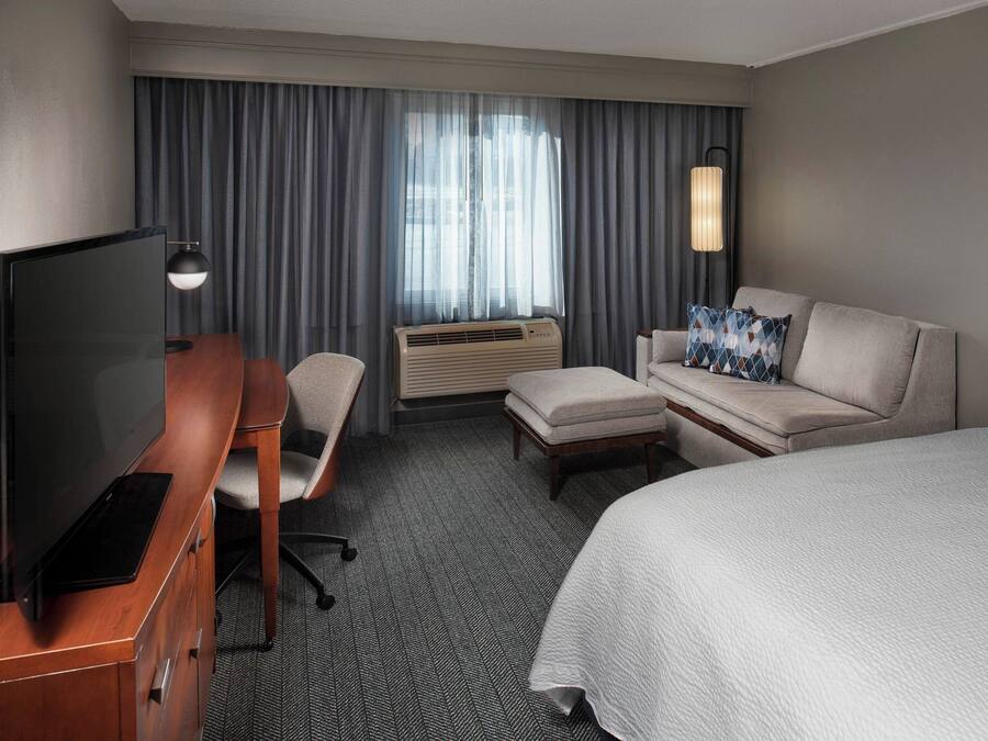 A hotel room with a bed, a desk with a TV, a chair, a sofa, and a window with curtains.