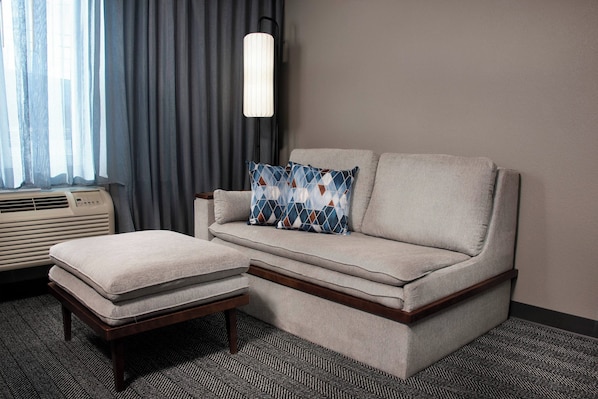 A modern hotel room with a sofa and ottoman, a standing lamp, and a window with curtains. - Courtyard by Marriott Winston-Salem University (Winston-Salem)