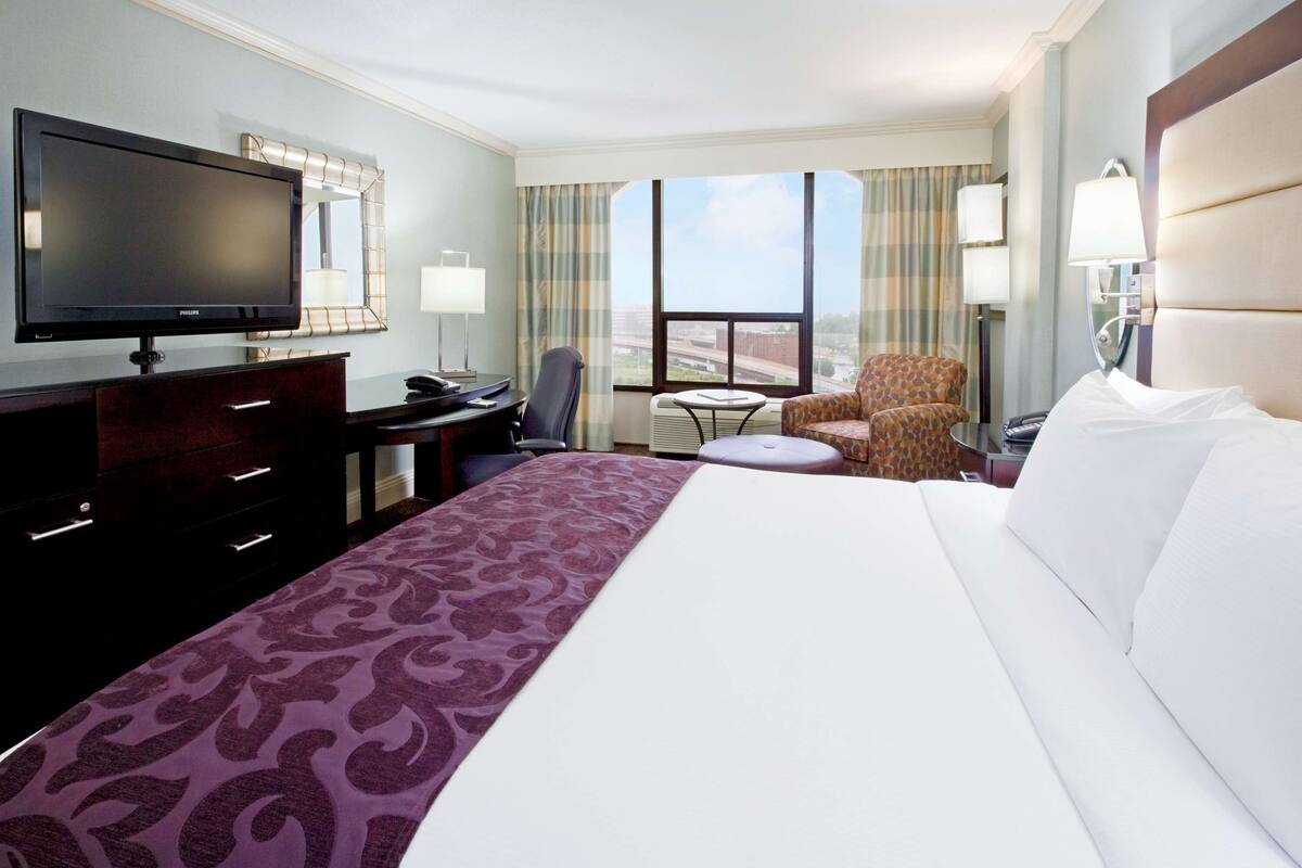A hotel room with a large bed, a desk with a flat-screen TV, a chair, a small table, and a view of the outdoors through large windows.
