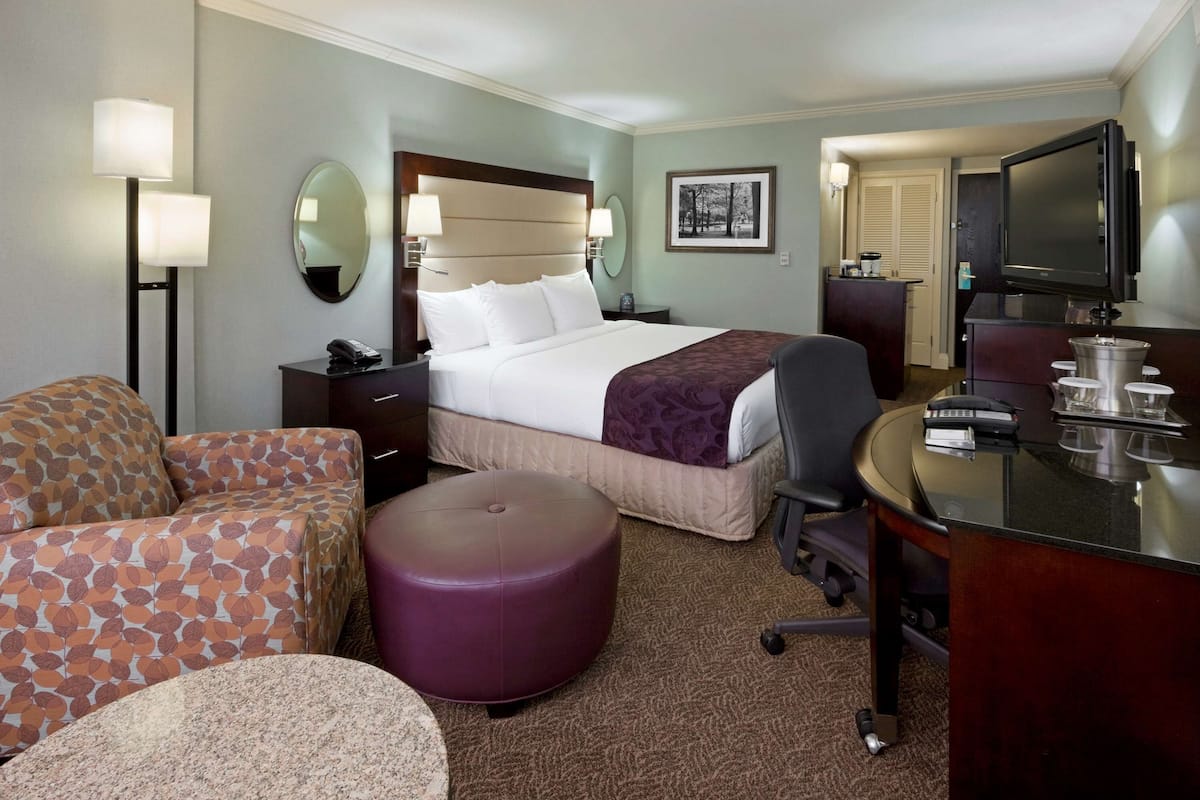 A hotel room with a bed, a desk, a chair, a sofa, a round ottoman, a TV, and a mirror.