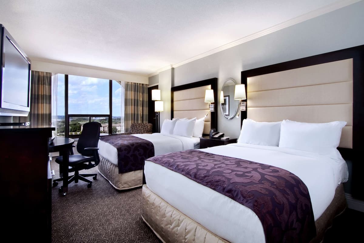 A hotel room with two beds, a desk, a chair, a TV, and a large window with a view.