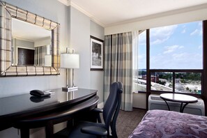 A hotel room with a bed, a desk with a phone, a chair, a small table, a lamp, a mirror, a picture on the wall, and a view of the city through large windows.