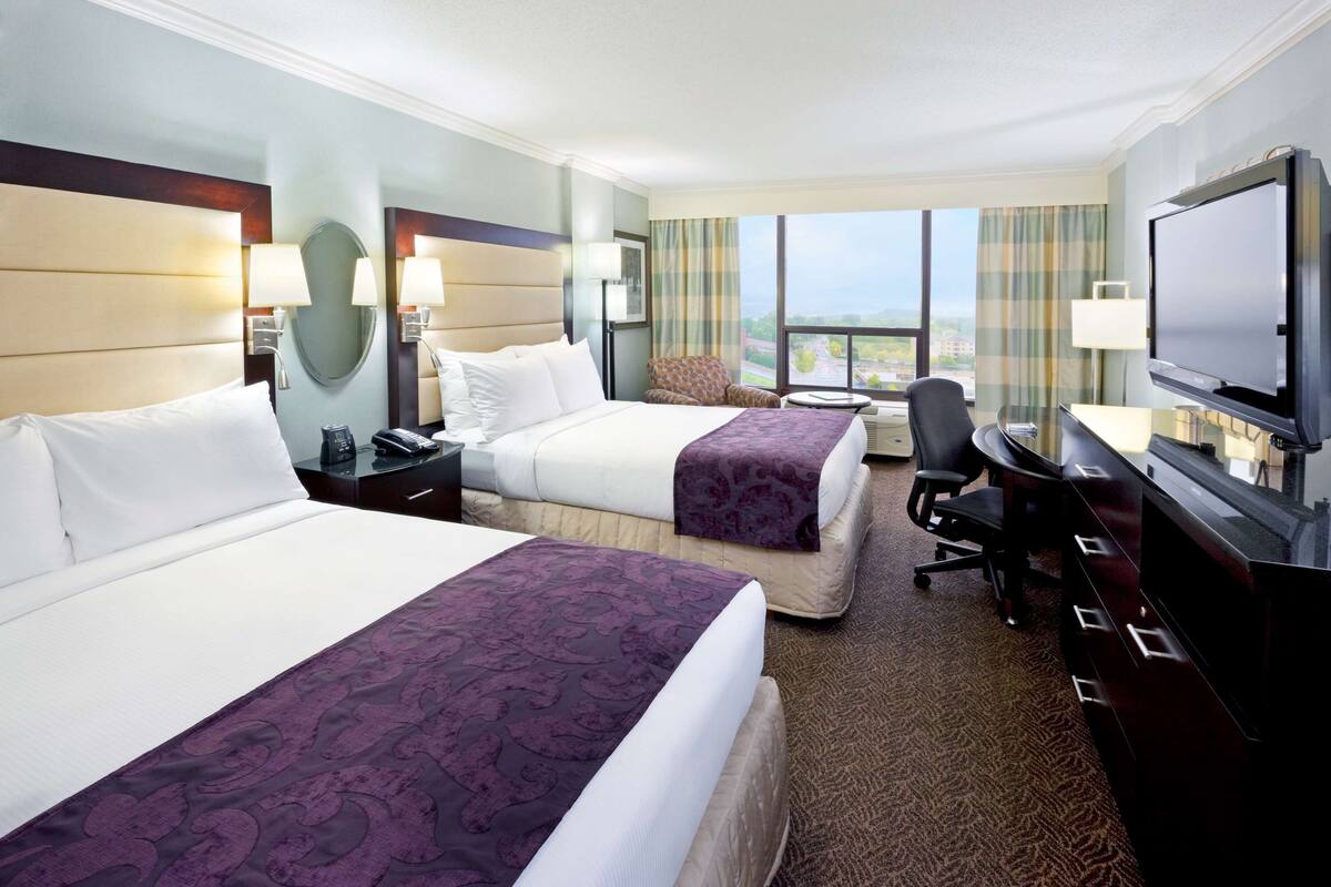 A hotel room with two beds, a desk, a chair, a television, and a large window with a view.