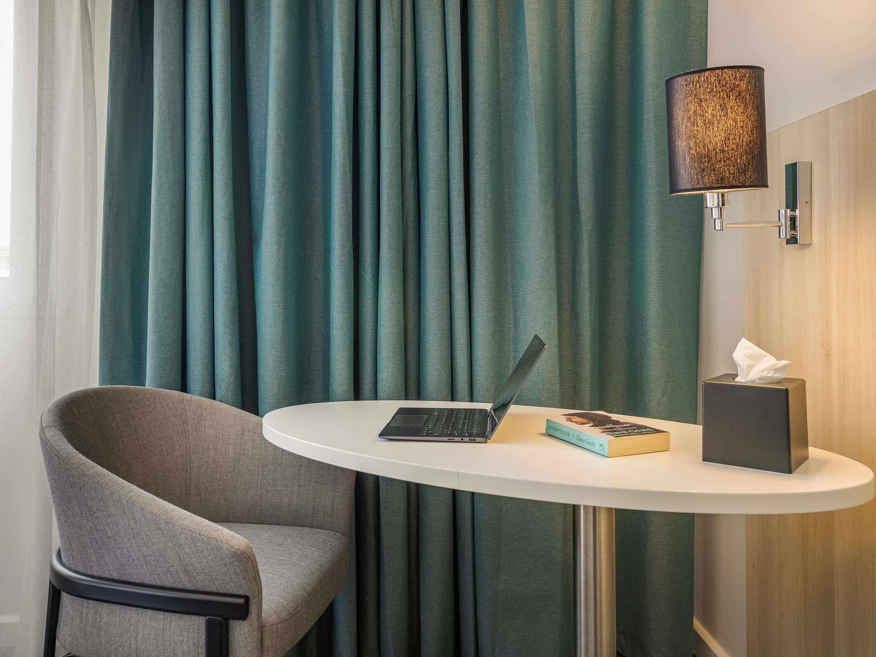 Habitación de hotel con cortinas azul verdoso, una computadora portátil sobre una mesita, un libro y una caja de pañuelos.