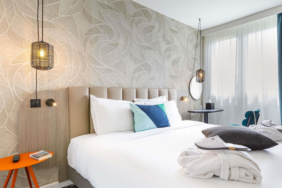 Una habitación de hotel con una cama grande, una mesita de noche con un control remoto, una pequeña mesa con un libro y un espejo en la pared.