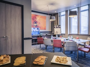 A hotel meeting room with tables, chairs, a large screen, and a painting on the wall.