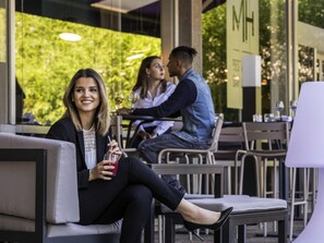 An outdoor seating area with tables and chairs, a glass wall with greenery, and a sign with 'MH' on it.