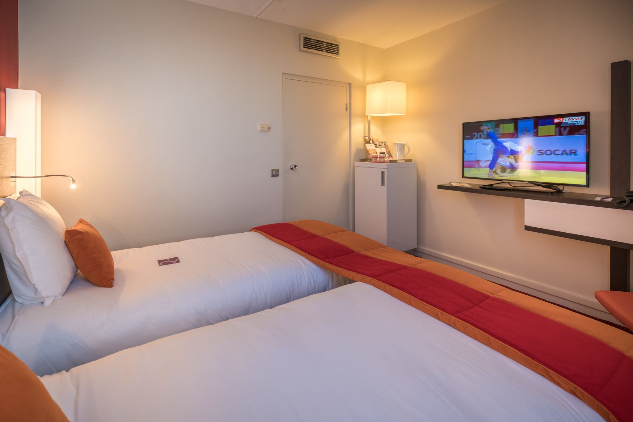 Habitación de hotel con dos camas, un televisor montado en la pared, un armario pequeño y una lámpara.