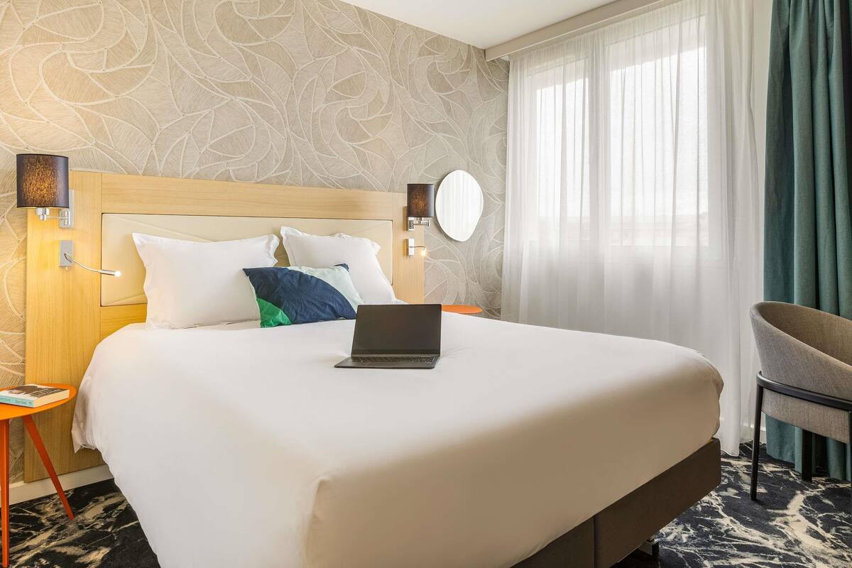 Habitación de hotel con una cama grande, una laptop sobre la cama, un espejo redondo y una silla.