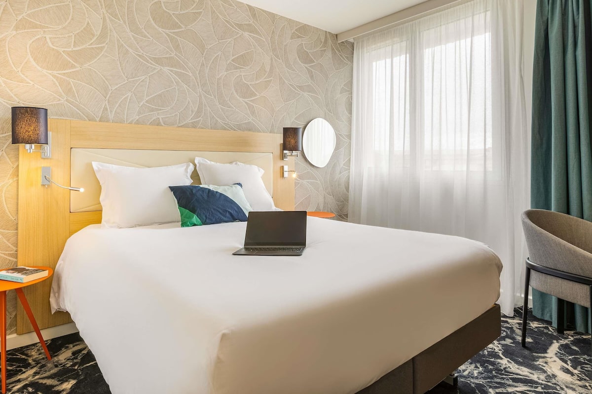 A hotel room with a large bed, a laptop on the bed, a round mirror, and a chair.